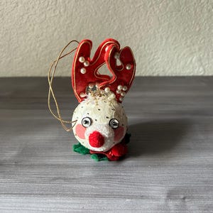 May include: A festive Christmas ornament shaped like a reindeer. The reindeer has a white sequined head, red antlers adorned with pearls, and a red nose. It has a gold string for hanging and green and red accents.