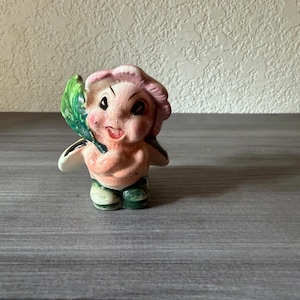 May include: A vintage ceramic figurine of a smiling pixie-like character. The figurine has a pink face and hair, a peach-colored torso, and green pants. It holds a green leaf and has white wings.