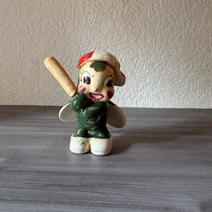 May include: A vintage ceramic figurine of a baseball player. The figurine is painted with a green uniform, white shoes, and a red and white cap. It is holding a tan baseball bat and has a smiling expression.