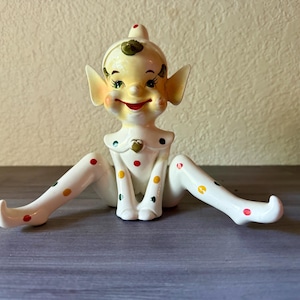 May include: A vintage ceramic elf figurine with a smiling face, large ears, and a pointed hat. The elf is white with colorful polka dots on the legs and chest. The figurine is sitting with legs spread, arms forward, and a heart on the chest.