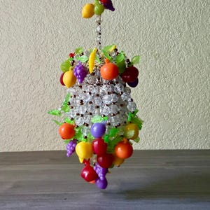 May include: A hanging decorative ornament crafted from clear faceted beads arranged in a cluster shape, adorned with small colorful plastic fruit charms including grapes, oranges, lemons, and bananas, interspersed with bright green plastic leaves.