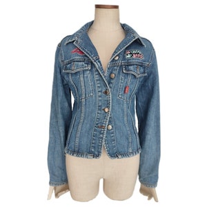 以下が含まれることがあります： 左側に赤と白のパッチ、右側にピンク、青、白のパッチが付いた青いデニムジャケット。ジャケットはボタン留め、長袖です。