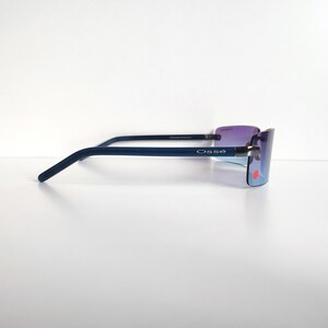 May include: Rimless sunglasses with a dark blue frame and purple-tinted lenses. The brand name "Oss&eacute;" is printed on the temple. The lenses have a gradient effect, transitioning from purple to blue. The sunglasses are set against a white background.