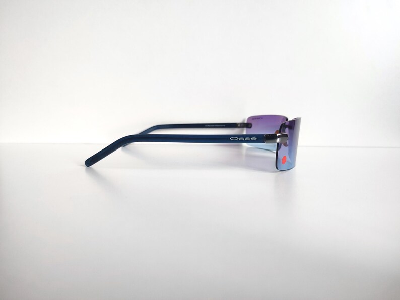 May include: Rimless sunglasses with a dark blue frame and purple-tinted lenses. The brand name "Oss&eacute;" is printed on the temple. The lenses have a gradient effect, transitioning from purple to blue. The sunglasses are set against a white background.
