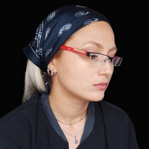 Könnte beinhalten: Ein dunkelblaues Kopftuch mit weißem Paisleymuster, kombiniert mit einer Halbrandbrille mit roten Bügeln und silbernen Akzenten. Die Brille hat klare Gläser und einen dünnen Metallrahmen, ergänzt durch silberne Creolen als Ohrschmuck.