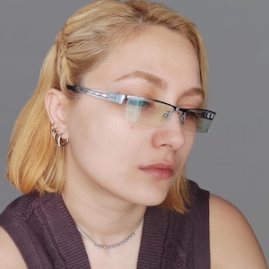 May include: A woman with short blonde hair wearing silver hoop earrings and a silver necklace. She is wearing a brown sleeveless top and glasses with a silver frame.