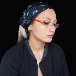 Könnte beinhalten: Eine Person trägt eine Brille mit einem rot-schwarzen Rahmen. Die Person trägt auch ein schwarzes Bandana mit weißem Paisley-Muster, eine schwarze Jacke und eine silberne Halskette mit Anhänger. Der Hintergrund ist schwarz.