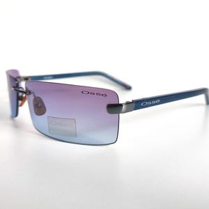 May include: Rimless sunglasses with a gradient lens, shifting from purple to blue. The frames are a dark blue colour, with the brand name "Oss&eacute;" printed on the temples. The sunglasses are set against a white background.