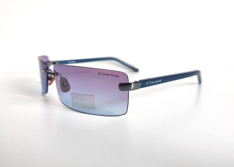 May include: Rimless sunglasses with a gradient lens, shifting from purple to blue. The frames are a dark blue colour, with the brand name "Oss&eacute;" printed on the temples. The sunglasses are set against a white background.