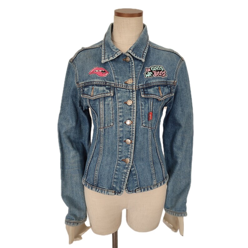 Pu&ograve; includere: Una giacca di jeans blu con una toppa a forma di labbra rosa e bianca e una toppa rosa e verde "In Girls We Trust".
