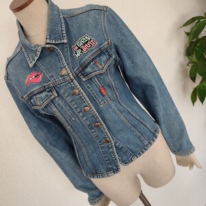 Pu&ograve; includere: Una giacca di jeans blu con toppe. Una toppa dice "In girls we trust" in lettere rosa e verdi. Un'altra toppa &egrave; una labbra rosse.