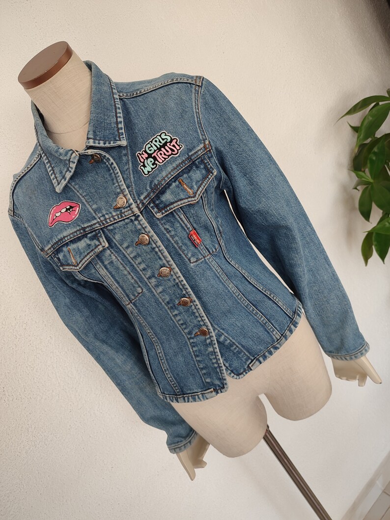 Pu&ograve; includere: Una giacca di jeans blu con toppe. Una toppa dice "In girls we trust" in lettere rosa e verdi. Un'altra toppa &egrave; una labbra rosse.