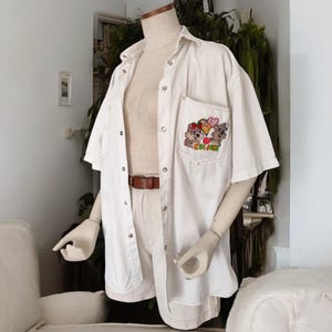 Può includere: Camicia bianca a maniche corte con bottoni e un disegno di cartoni animati ricamato sulla tasca. Il disegno presenta tre animali dei cartoni animati con un cuore e il testo "Go For It!"