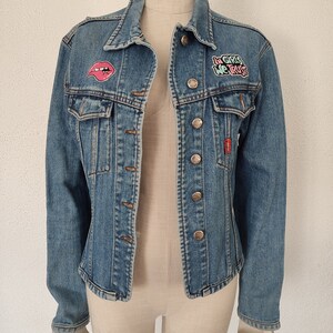 Pu&ograve; includere: Una giacca di jeans blu con una toppa rosa e verde che dice "In Girls We Trust" e una toppa di labbra rosa.
