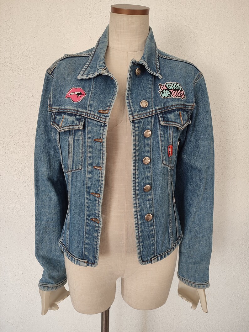 Pu&ograve; includere: Una giacca di jeans blu con una toppa rosa e verde che dice "In Girls We Trust" e una toppa di labbra rosa.