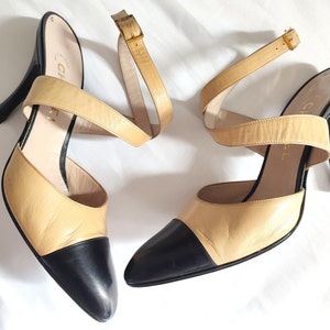 May include: A pair of beige leather heeled sandals with black leather toe caps and ankle straps. The sandals have a gold buckle on the ankle strap.