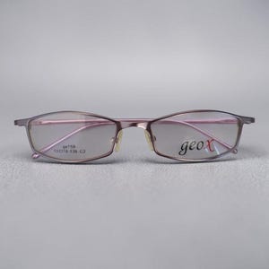 Può includere: Un paio di occhiali metallici color malva con montatura sottile. La lente sinistra presenta il testo "gx159 53 18-135 C2" e la lente destra mostra il marchio "geox" in un carattere corsivo nero. Gli occhiali hanno una forma rettangolare.