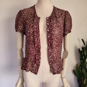 May include: A maroon sheer blouse with a white floral pattern. The blouse has a button-up front and short sleeves with ruffles at the hem.