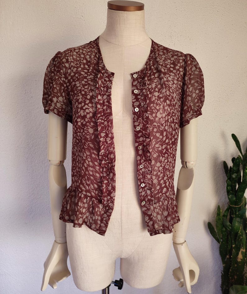 May include: A maroon sheer blouse with a white floral pattern. The blouse has a button-up front and short sleeves with ruffles at the hem.