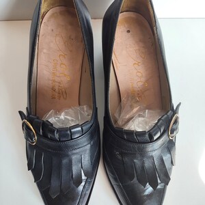 May include: A pair of black leather pumps with pointed toes and fringe detailing. Each shoe features a gold-colored buckle and a high heel. The insoles are a light tan color with gold lettering. The shoes are vintage.