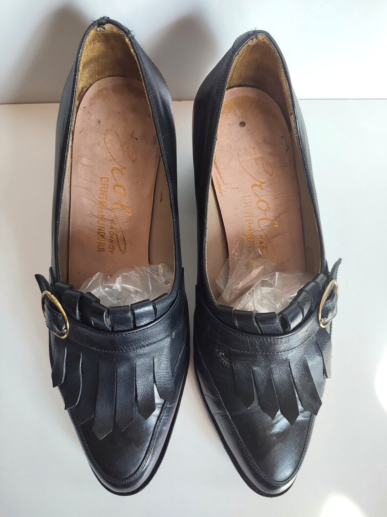 May include: A pair of black leather pumps with pointed toes and fringe detailing. Each shoe features a gold-colored buckle and a high heel. The insoles are a light tan color with gold lettering. The shoes are vintage.