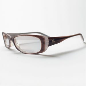 Op de afbeelding: Bruine rechthoekige bril met een gradiëntontwerp op de monturen. De veren zijn donkerder bruin met een gouden accent. De lenzen zijn helder.