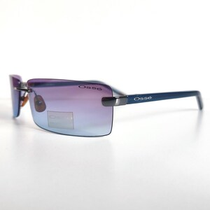 May include: Rimless sunglasses with gradient purple and blue lenses. The frames are a dark blue colour, with the brand name "Oss&eacute;" printed on the temples and lenses. The sunglasses are set against a white background.