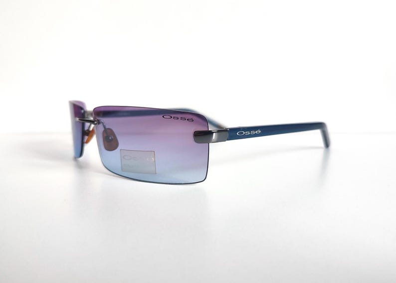 May include: Rimless sunglasses with gradient purple and blue lenses. The frames are a dark blue colour, with the brand name "Oss&eacute;" printed on the temples and lenses. The sunglasses are set against a white background.
