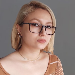 Pode incluir: Uma mulher com cabelo loiro vestindo uma camisa listrada marrom e branca, um colar de ouro e óculos de armação preta. Ela está olhando diretamente para a câmera.