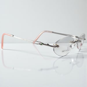 May include: Rimless spectacles with oval lenses and silver frames. The arms have a pink hue at the ends and a decorative element with a small green accent. The text "G427 510" is printed on the lens.