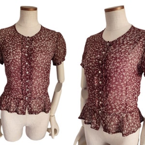 May include: A maroon sheer blouse with a white floral pattern and a button-up front. The blouse has a ruffled hem and short sleeves.