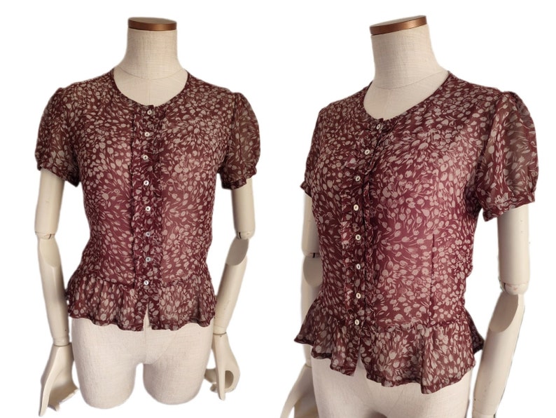 May include: A maroon sheer blouse with a white floral pattern and a button-up front. The blouse has a ruffled hem and short sleeves.