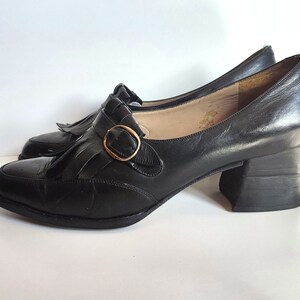 May include: A pair of black leather Mary Jane style shoes with a block heel. The shoes feature a decorative fringe detail and a gold-colored buckle closure. The heels are approximately 2 inches high.