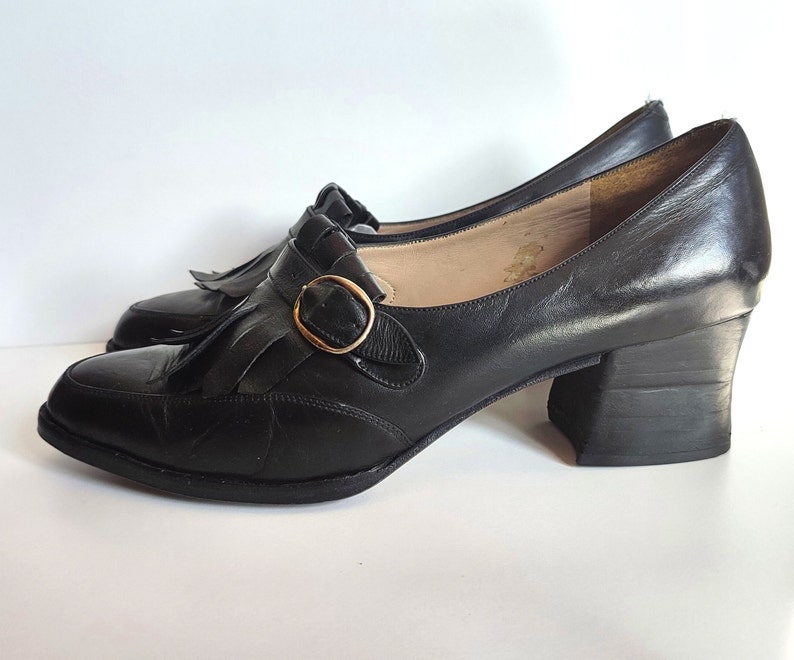 May include: A pair of black leather Mary Jane style shoes with a block heel. The shoes feature a decorative fringe detail and a gold-colored buckle closure. The heels are approximately 2 inches high.