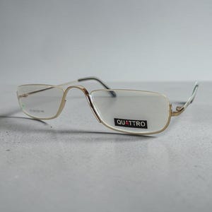 Può includere: Occhiali in metallo color oro con montatura semi-cerchiata. Le lenti sono trasparenti e la lente destra presenta un adesivo nero con il testo bianco QUATTRO. Le aste sottili hanno punte scure e il ponte è arcuato per un aspetto classico.