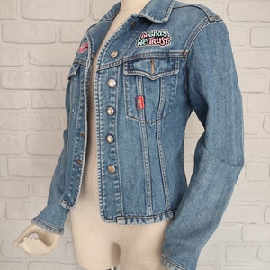 Pu&ograve; includere: Una giacca di jeans blu chiaro con una toppa che dice "In Girls We Trust" in lettere rosa e blu. La giacca ha una chiusura con bottoni e due tasche anteriori.