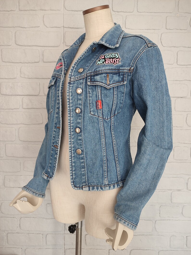 Pu&ograve; includere: Una giacca di jeans blu chiaro con una toppa che dice "In Girls We Trust" in lettere rosa e blu. La giacca ha una chiusura con bottoni e due tasche anteriori.