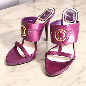May include: A pair of purple high-heeled sandals with gold buckles featuring the Christian Dior logo. The sandals have a platform sole and a thin strap across the toe.