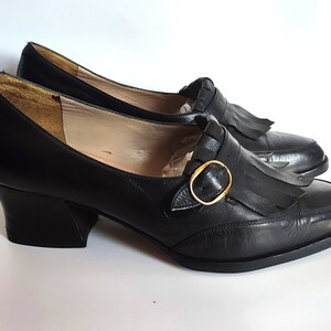 May include: A pair of black leather shoes with a low heel and a rounded toe. Each shoe features a decorative fringe and a gold-colored buckle. The shoes are displayed on a white surface.