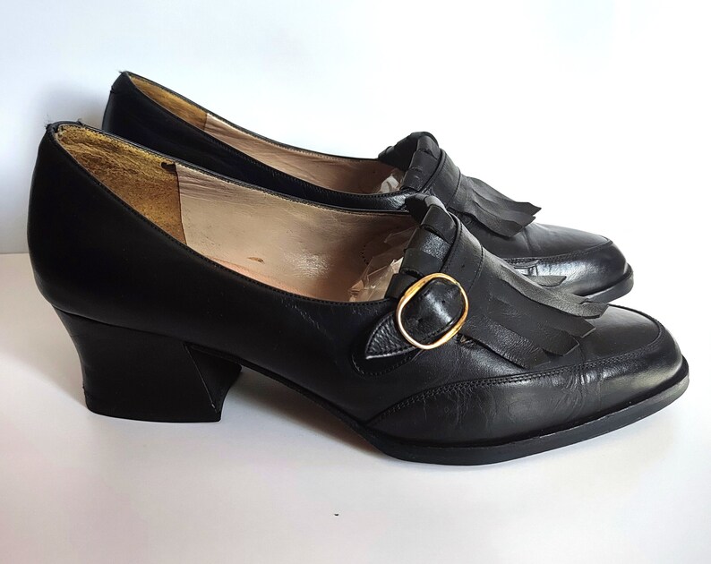 May include: A pair of black leather shoes with a low heel and a rounded toe. Each shoe features a decorative fringe and a gold-colored buckle. The shoes are displayed on a white surface.