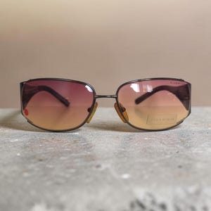 Könnte beinhalten: Sonnenbrille mit einem dunklen Metallrahmen und Verlaufsgläsern, die von einem tiefen Violett oben zu einem warmen Bernstein unten übergehen. Das rechte Glas hat einen kleinen roten Punkt und das linke Glas einen rechteckigen Aufkleber mit dem Text CLUB ROMAN.