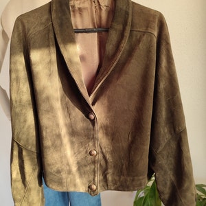 Puede incluir: Una chaqueta de ante verde con cuello y cierre de botones. La chaqueta tiene mangas largas y un corte holgado.
