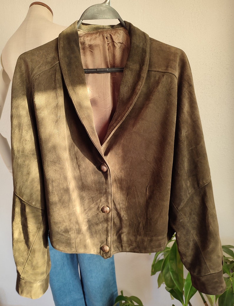 Puede incluir: Una chaqueta de ante verde con cuello y cierre de botones. La chaqueta tiene mangas largas y un corte holgado.