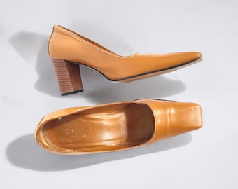 May include: A pair of tan leather heeled pumps with square toes. The shoes have a wooden heel and the insole is marked "Gucci Made in Italy."