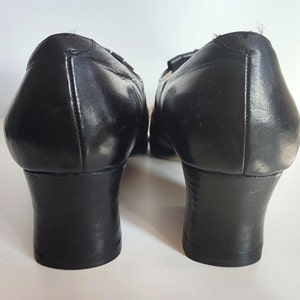 May include: Pair of black leather shoes with a low heel. The shoes have a rounded toe and a small decorative detail on the top. The heels are approximately 2 inches high.