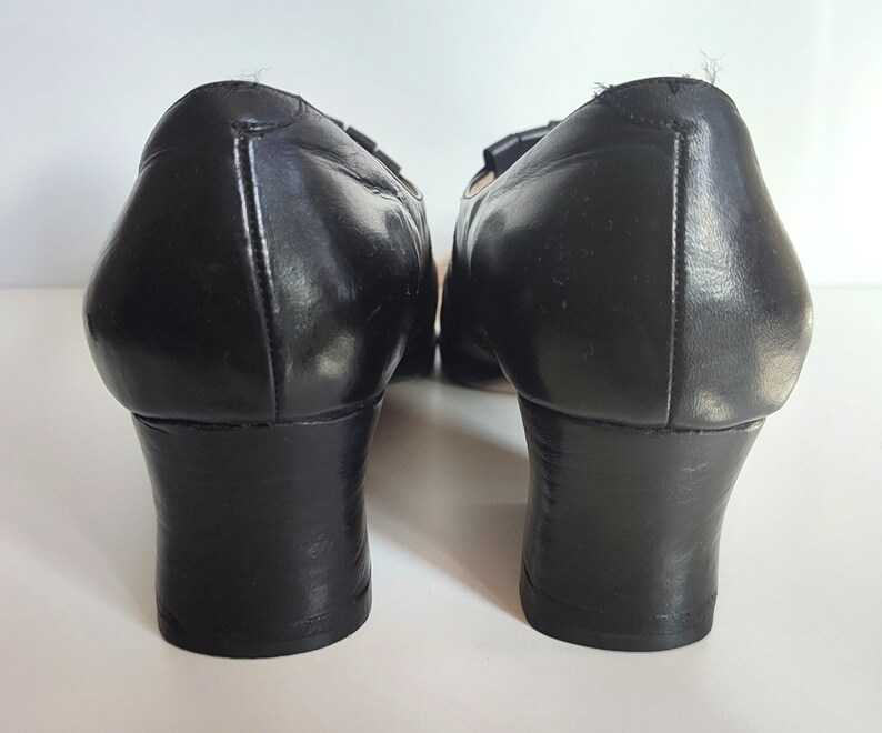 May include: Pair of black leather shoes with a low heel. The shoes have a rounded toe and a small decorative detail on the top. The heels are approximately 2 inches high.
