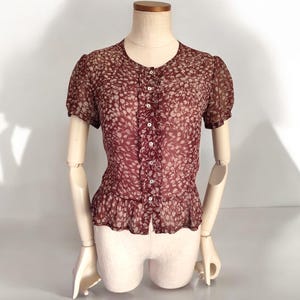 May include: A maroon sheer blouse with a white floral pattern and a button-up front. The blouse has short sleeves and a ruffled hem.