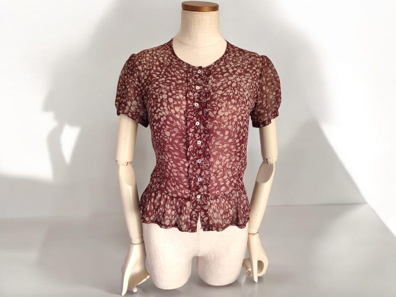 May include: A maroon sheer blouse with a white floral pattern and a button-up front. The blouse has short sleeves and a ruffled hem.