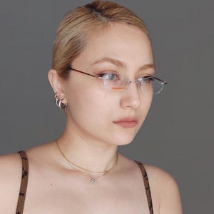 May include: Rimless eyeglasses with a thin, brown metal frame and clear lenses. The glasses have a rectangular shape and are worn by a person with blonde hair. The person is also wearing small silver hoop earrings and a gold necklace.