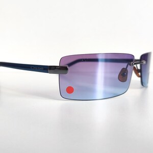 May include: Rimless sunglasses with gradient purple and blue lenses. The frames are a dark blue colour, with the brand name "Osse" printed on the temple. A small red dot is on the lower left lens.